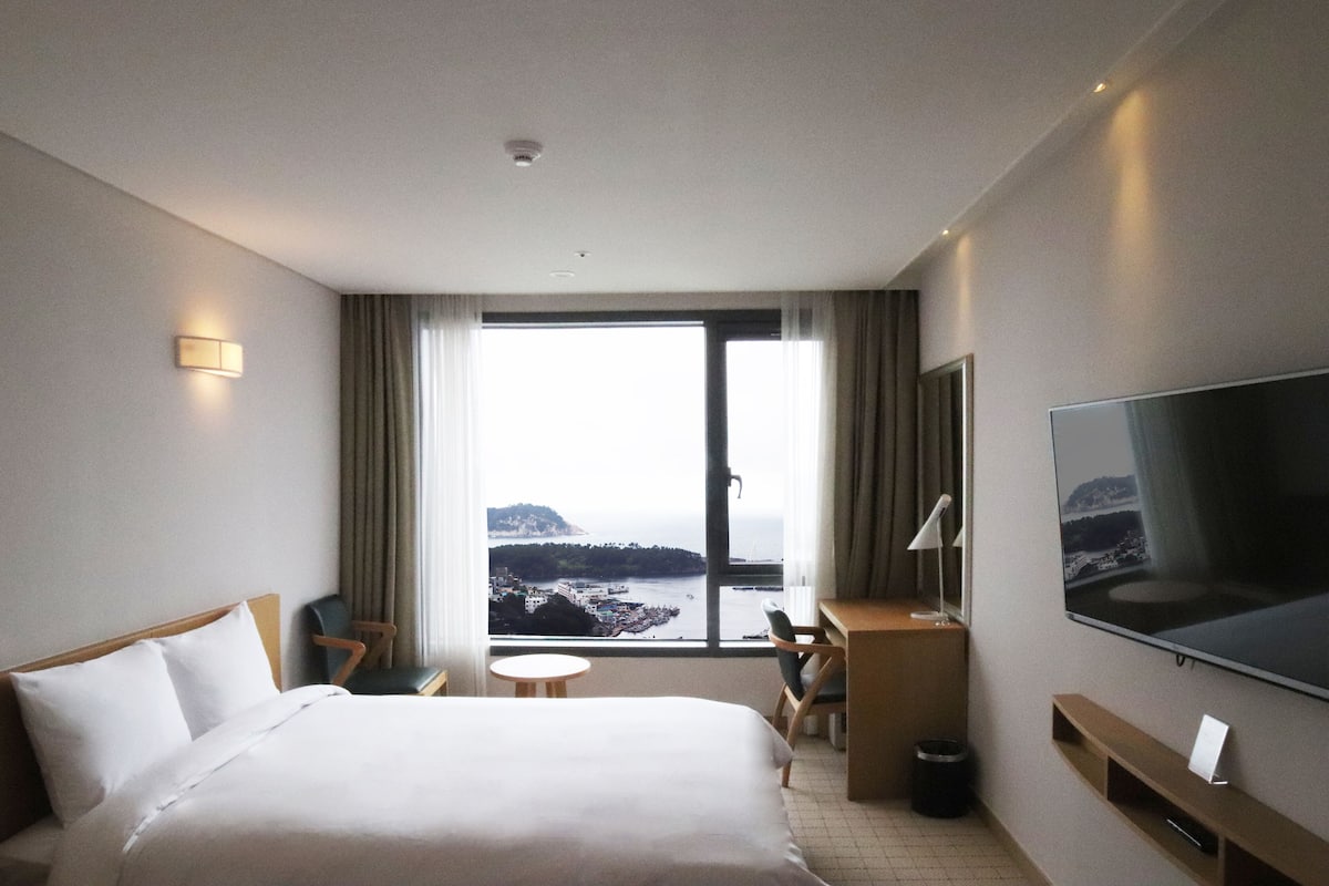 A hotel room with a bed, a desk, a chair, a TV, and a view of the sea.