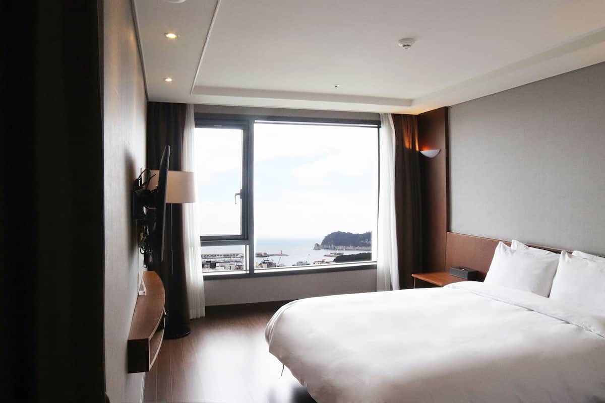 A hotel room with a large bed, a nightstand, a desk, and a window offering a view of the sea and a cliff.