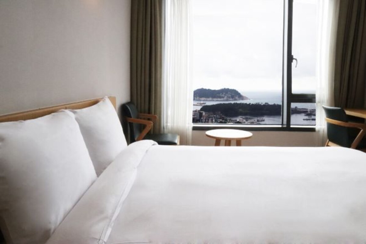 Quarto de hotel com uma cama, uma mesinha e vista para a paisagem costeira através de grandes janelas.