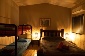 Ein Schlafsaalzimmer mit Etagenbetten, einem Einzelbett, zwei Nachttischlampen, einem gerahmten Bild an der Wand und einer Klimaanlage.