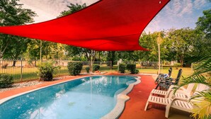 Una piscina con una vela sombreadora roja, rodeada por una cerca y algunas sillas y plantas cerca.