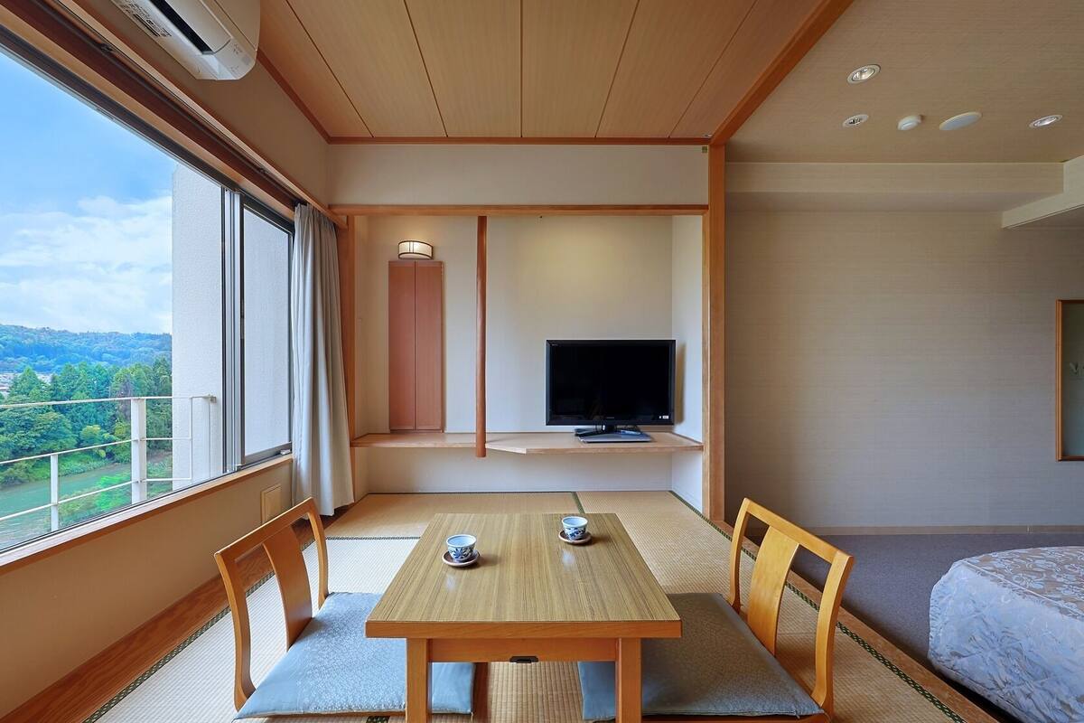 Habitación de hotel con una mesa de madera, sillas, televisor y un ventanal con vistas a un paisaje verde.