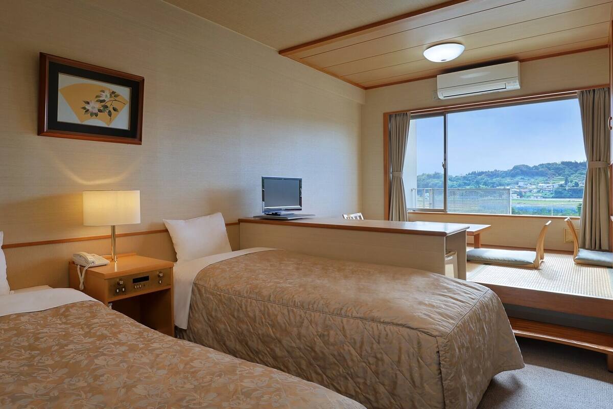 Habitación de hotel con dos camas, un escritorio con computadora, televisor y un amplio ventanal con vista a la ciudad.