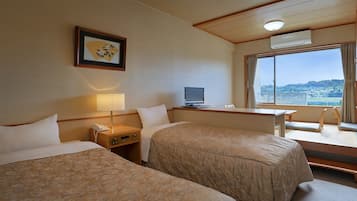 Ein Hotelzimmer mit zwei Betten, einem Schreibtisch mit Computer, einem Fernseher und einem großen Fenster mit Blick auf eine Stadtlandschaft.