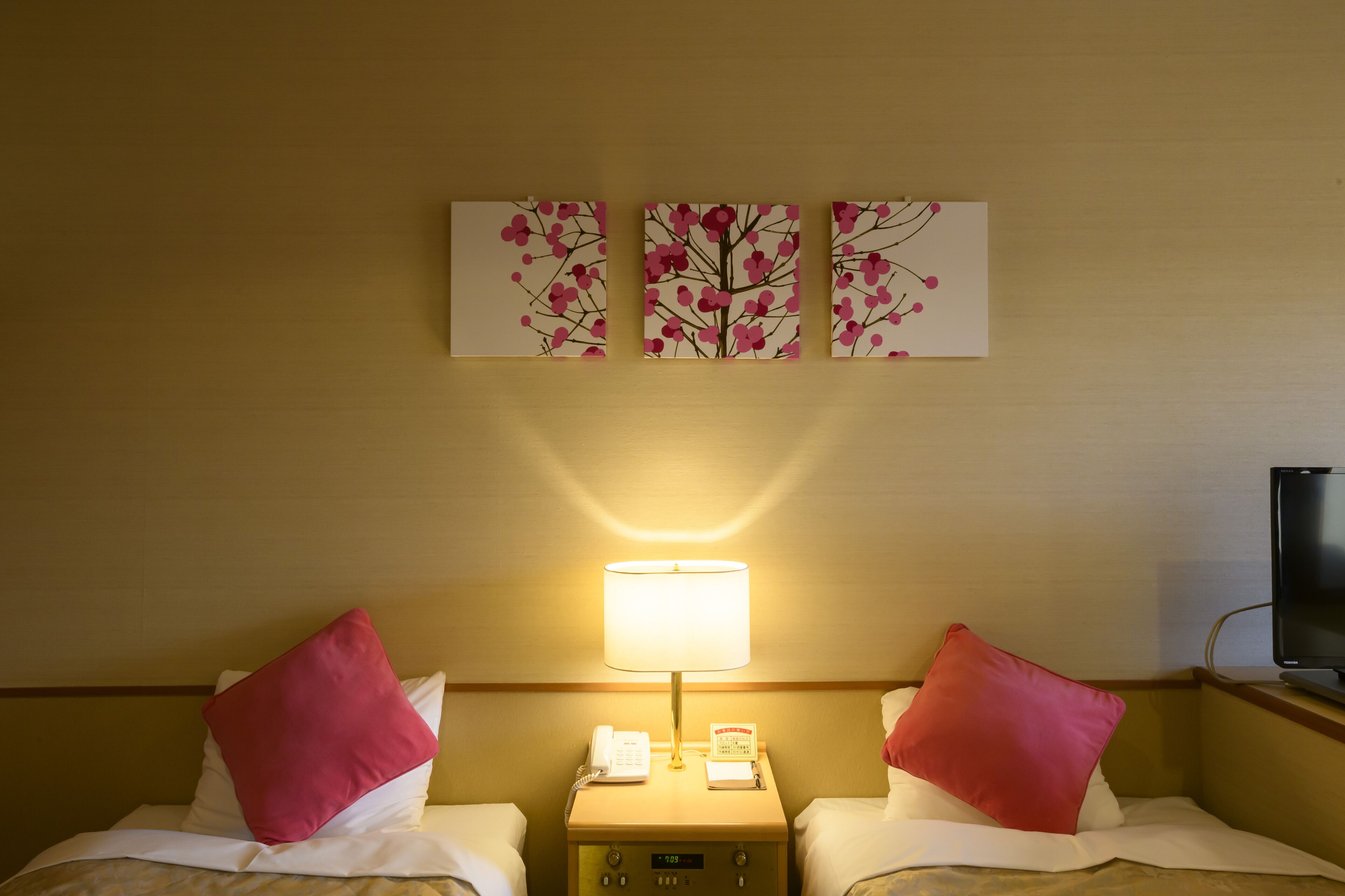 A hotel room with two beds, each with a red pillow, a bedside table with a lamp, a phone, and a small clock. There are three wall art pieces above the beds.