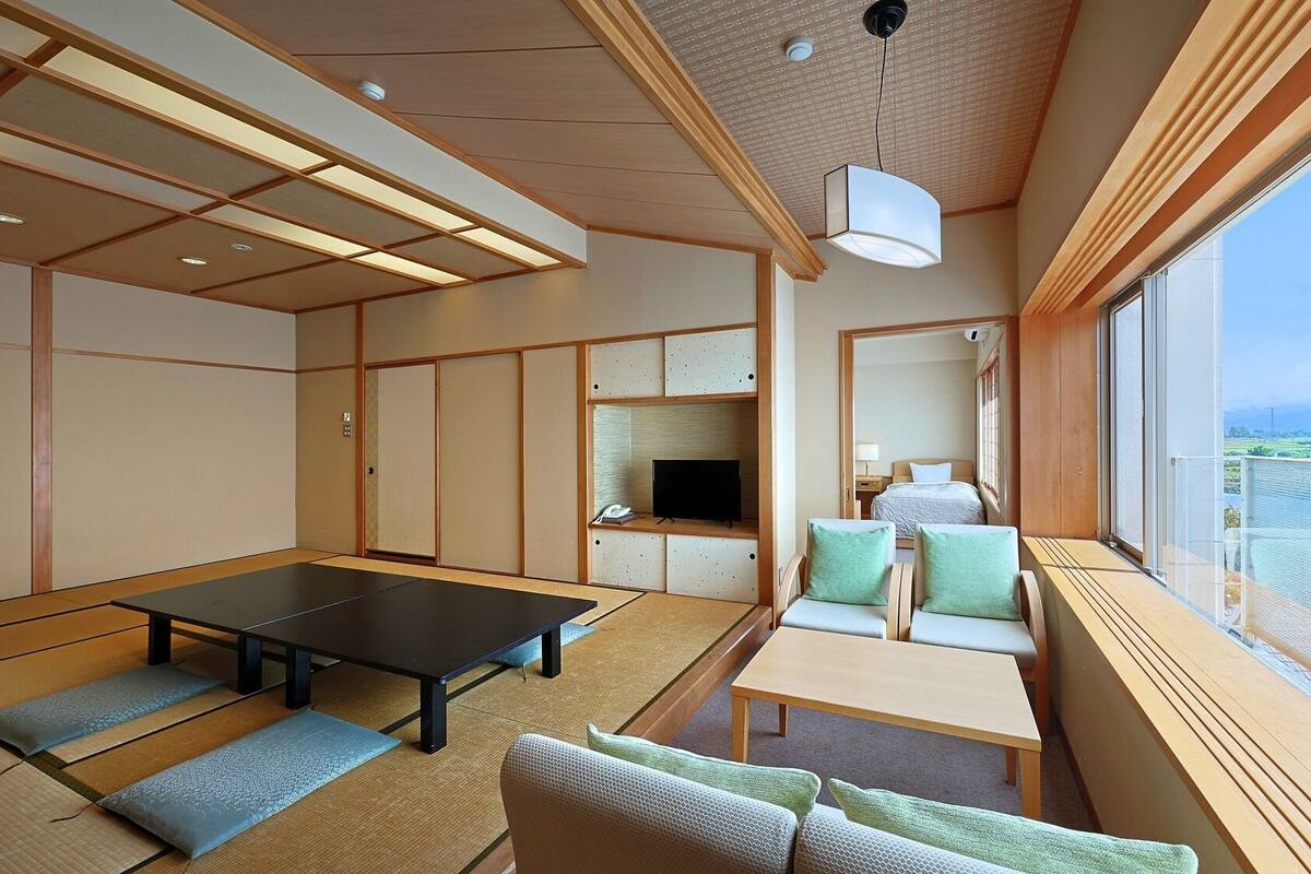 Una habitación japonesa tradicional con suelo de tatami, una mesa baja de tatami, un sofá con cojines, un televisor y un balcón con vistas.