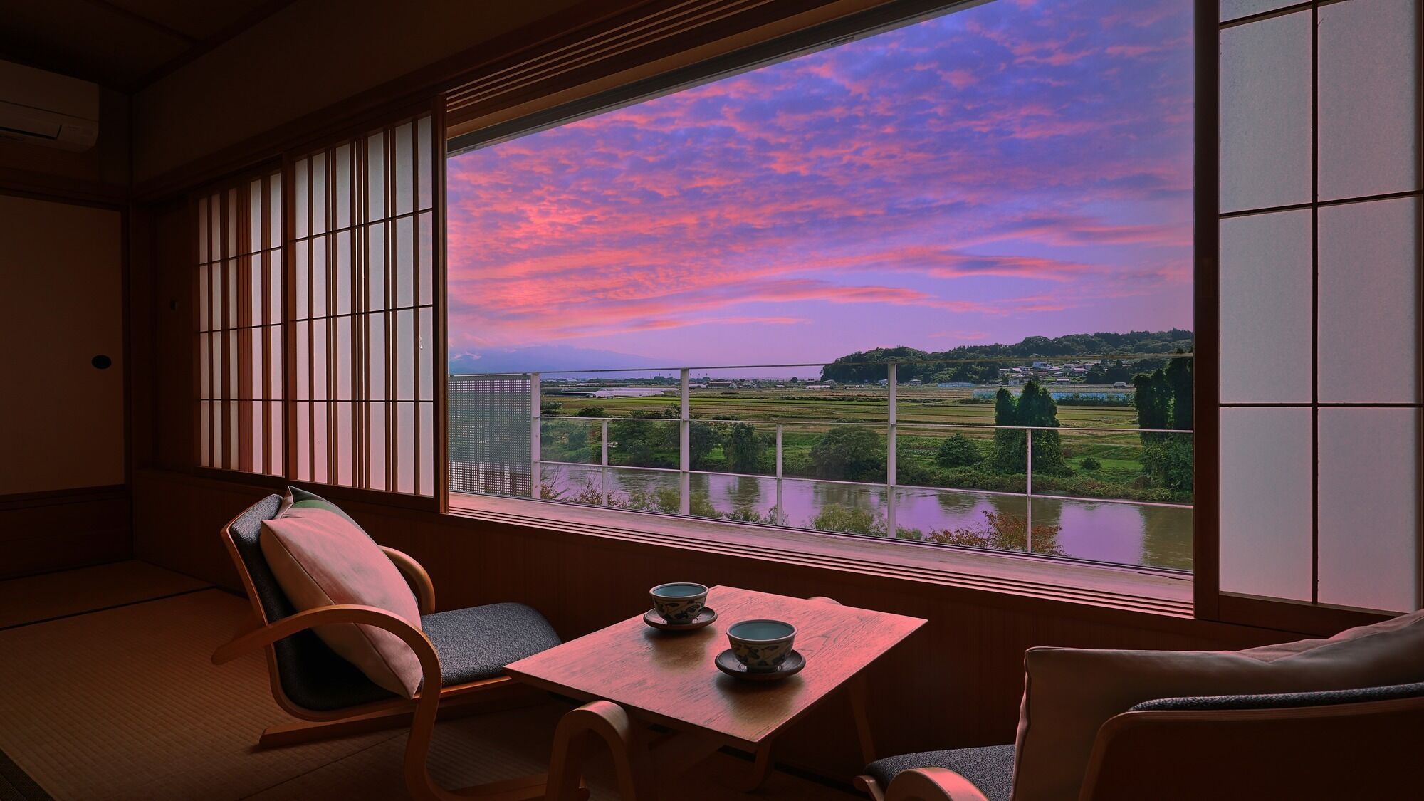 Une pièce au sol recouvert de tatamis, une petite table avec deux tasses, et une vue sur un paysage à travers un paravent en papier de riz.
