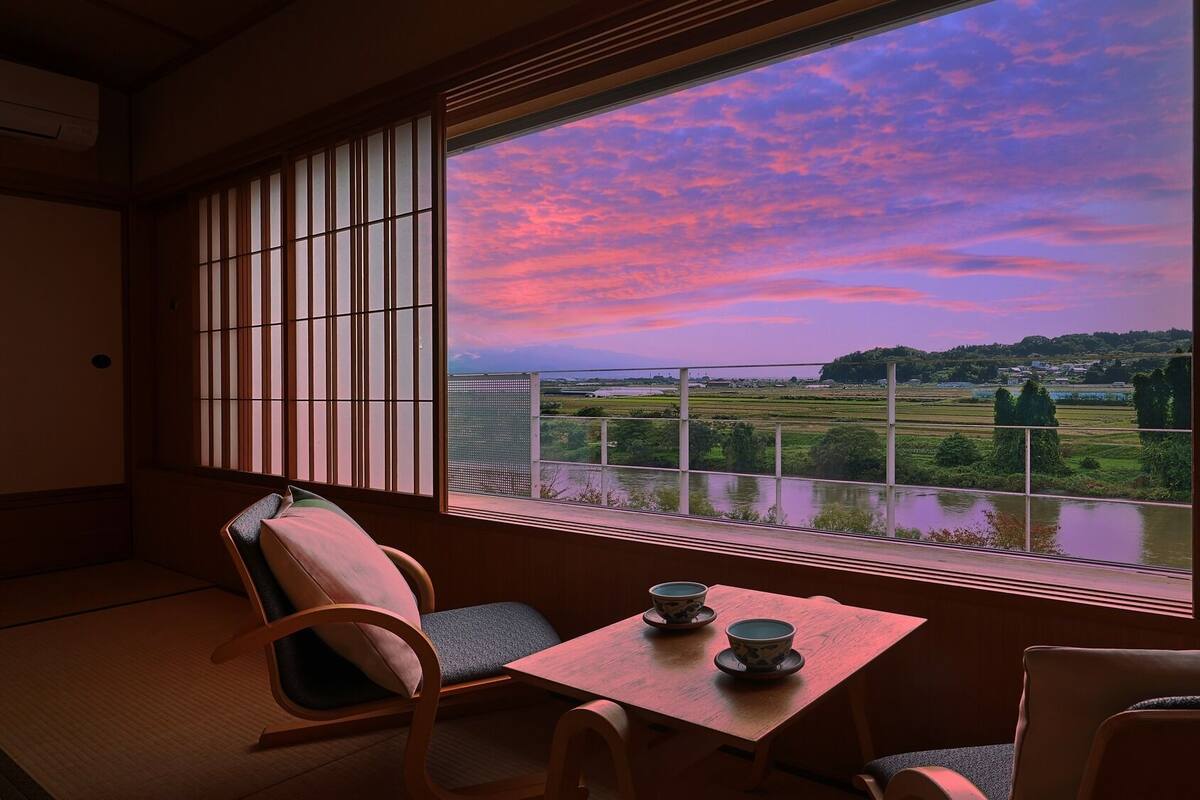 Habitación con suelo de tatami, una mesa baja con dos tazas y vista a un paisaje a través de un celosía japonesa.