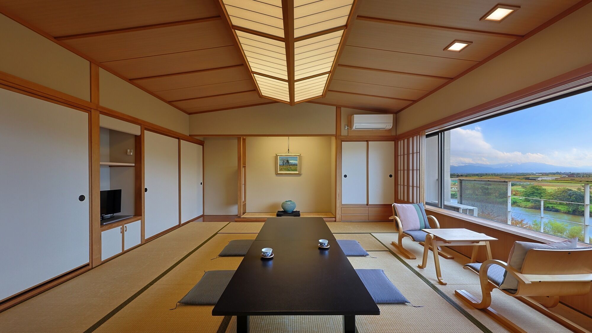 A traditional Japanese-style room with tatami flooring, a low table with cushions, a television, and a large window offering a view of a landscape.