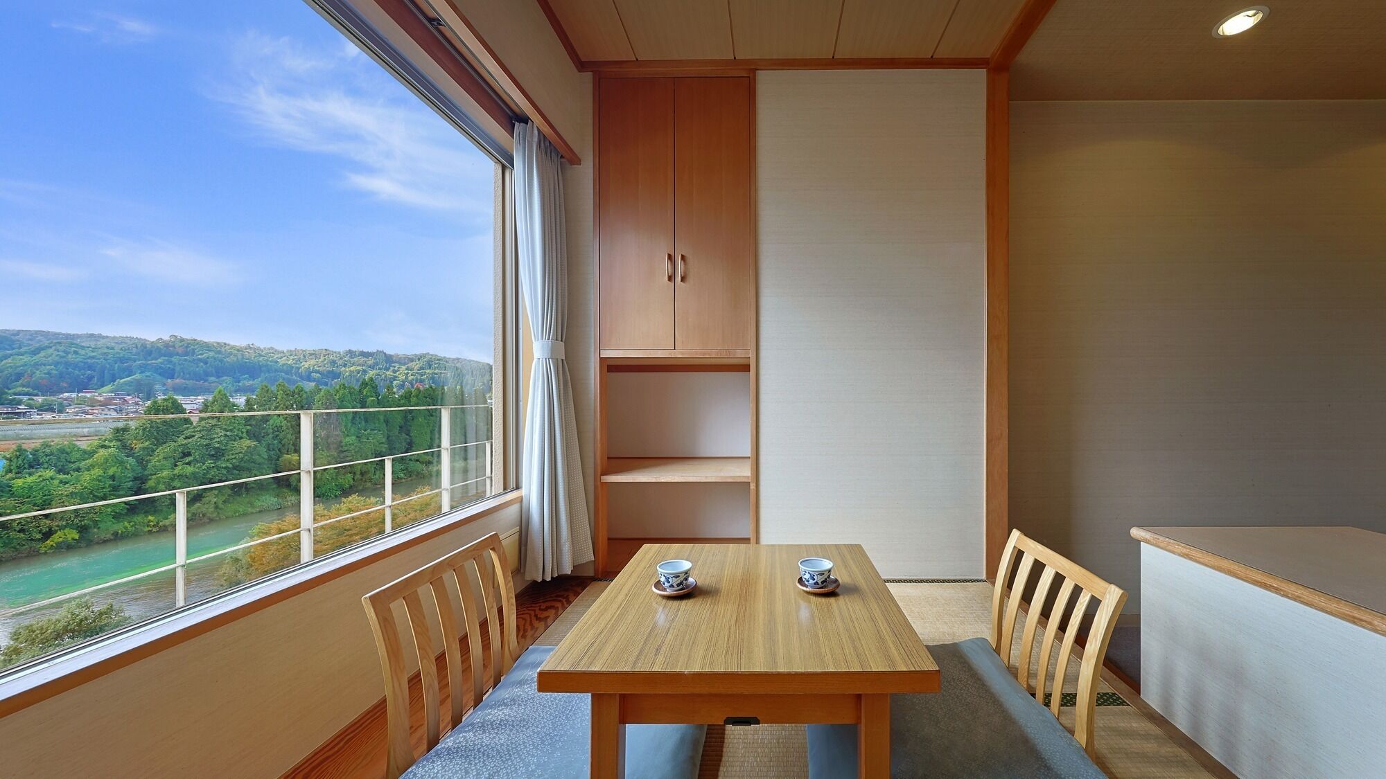 A room with a wooden table and chairs, a view of a river and hills, and a large window with curtains.
