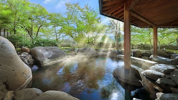 Un área tranquila de aguas termales al aire libre, rodeada de grandes rocas, un pabellón de madera y abundante vegetación en el fondo.