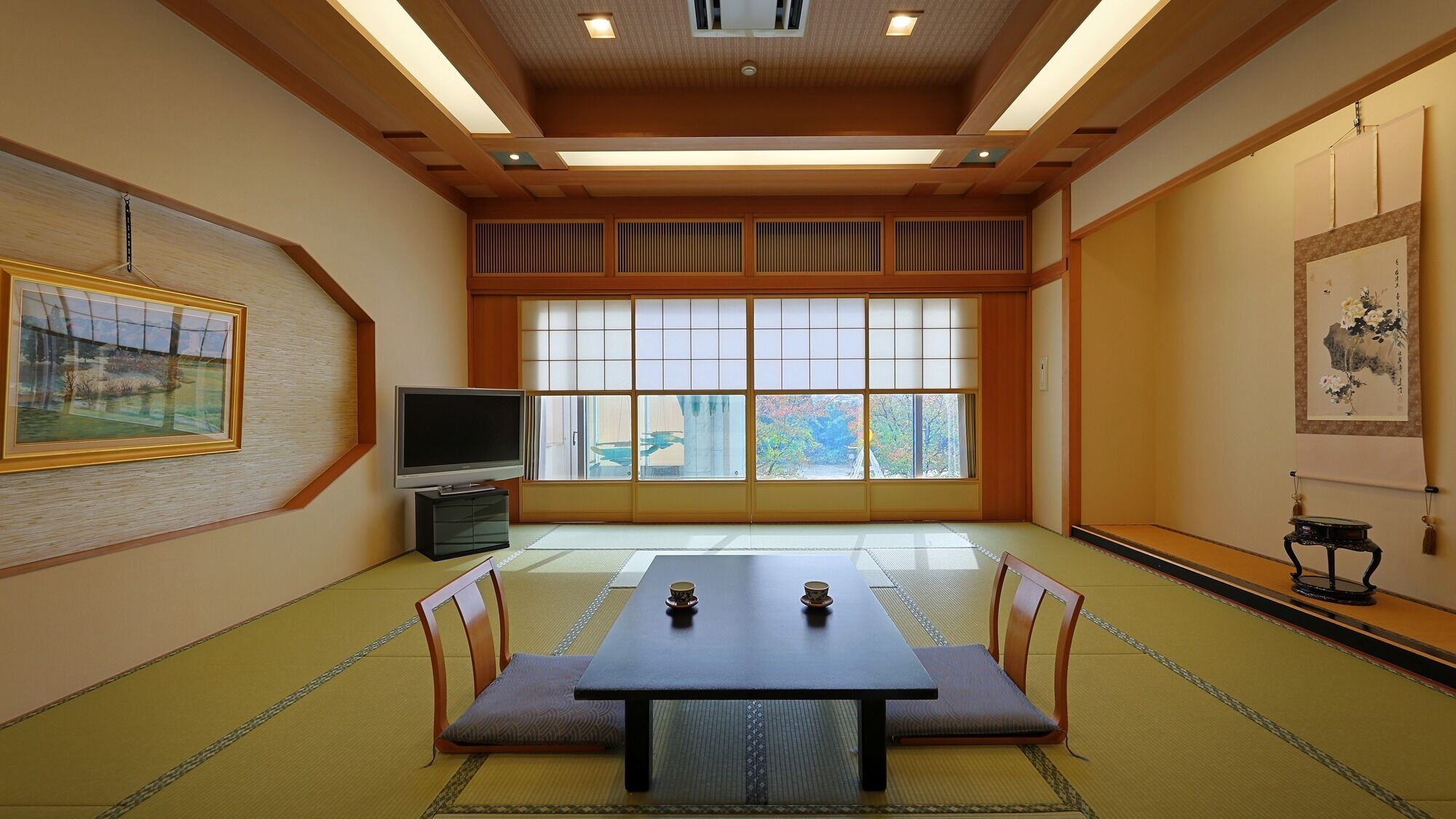 Une pièce de style japonais traditionnel, avec un sol recouvert de tatamis, une table basse avec des coussins, un téléviseur, des portes coulissantes en papier de riz (shoji) et des œuvres d’art accrochées aux murs.