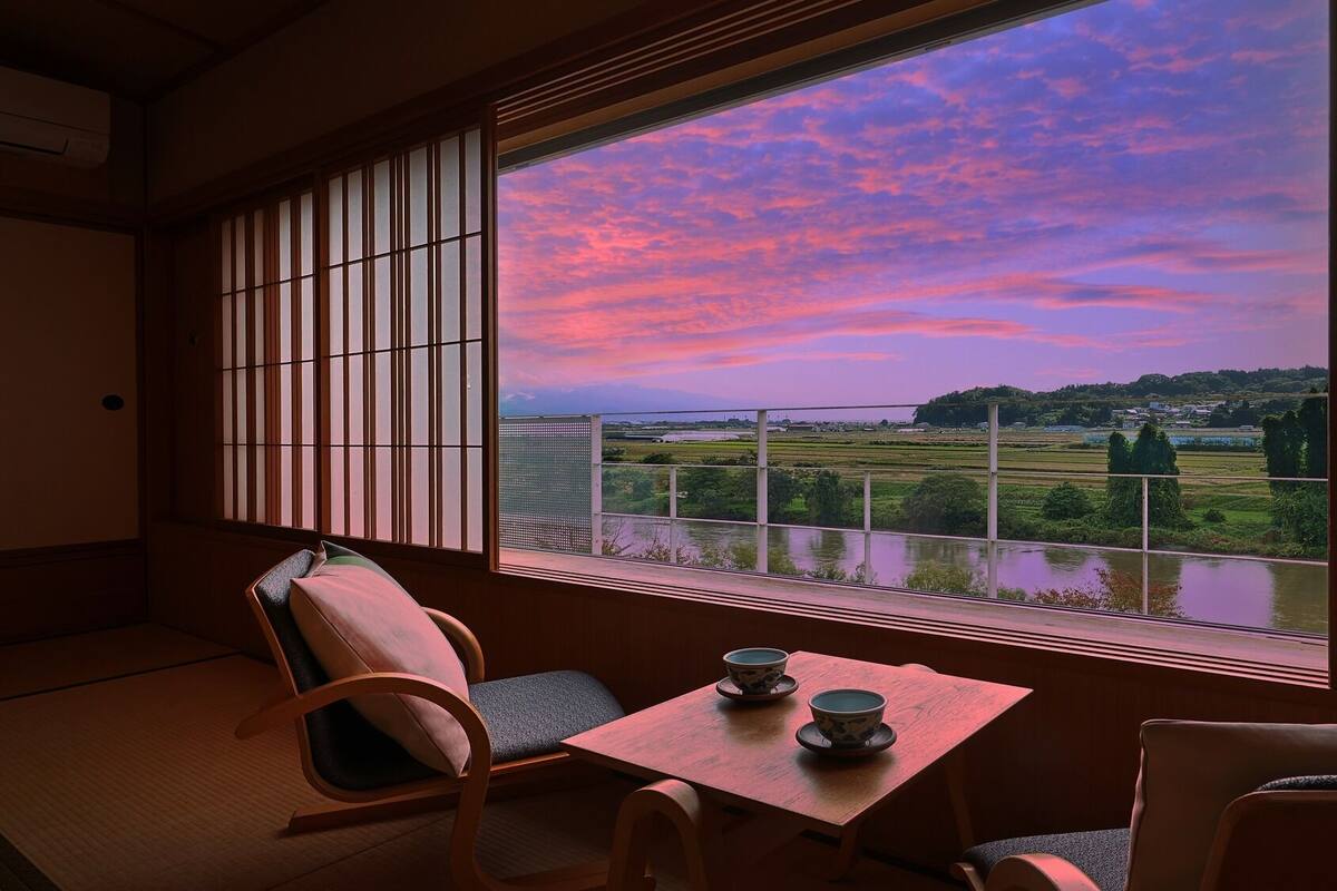 Una habitación japonesa tradicional con suelo de tatami, una mesa baja con dos cuencos y vistas a un paisaje al atardecer.