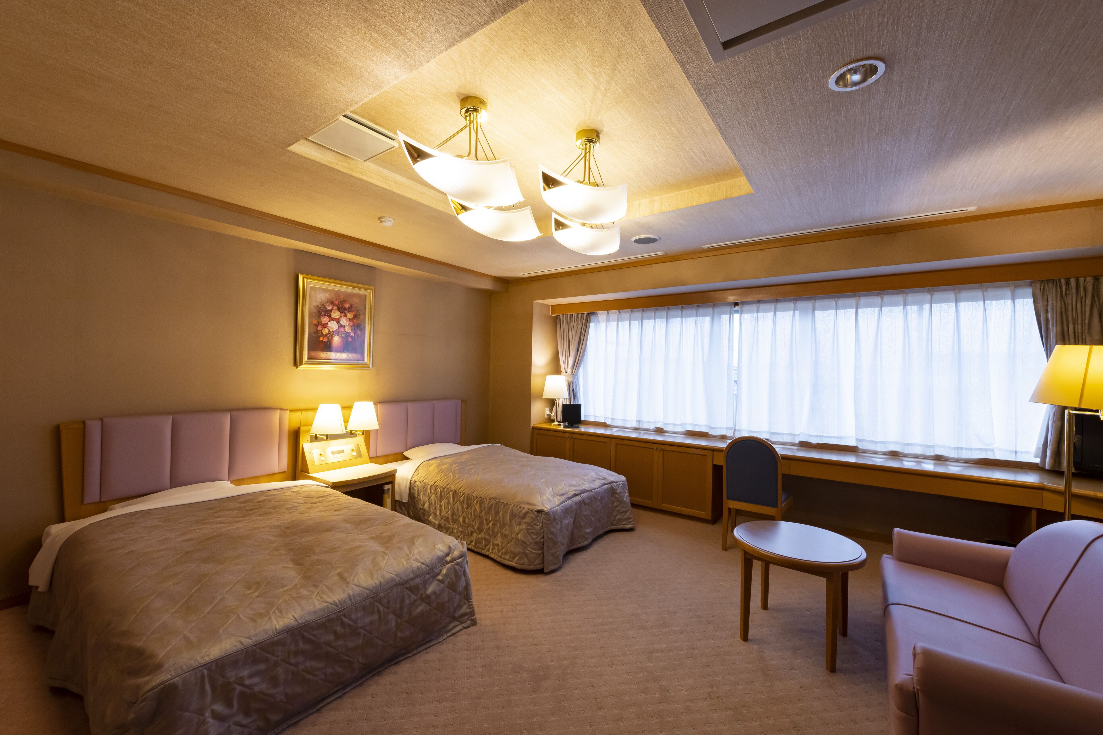 A hotel room with two beds, a desk, a chair, and a sofa. There is a window with curtains and a painting on the wall.