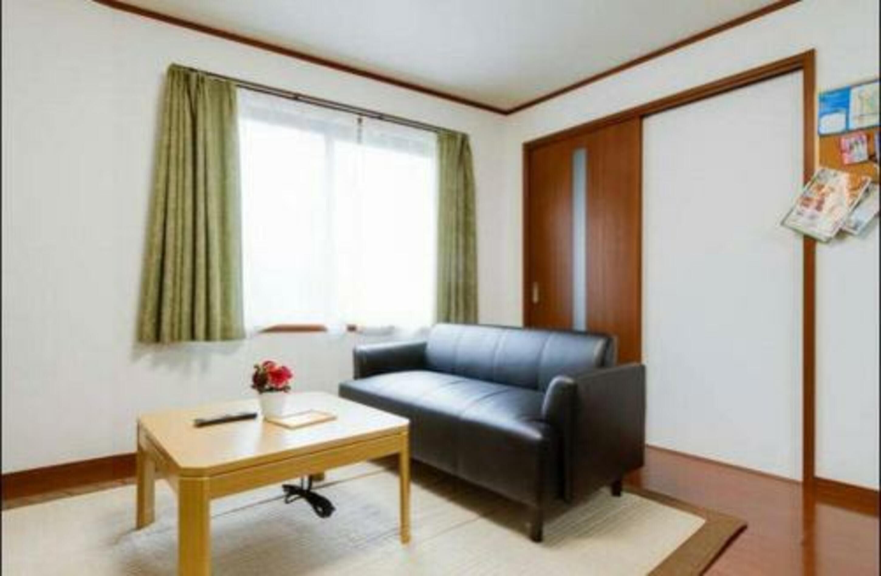 Una sala pequeña con un sofá de cuero negro, una mesa de centro de madera y una ventana con cortinas verdes.