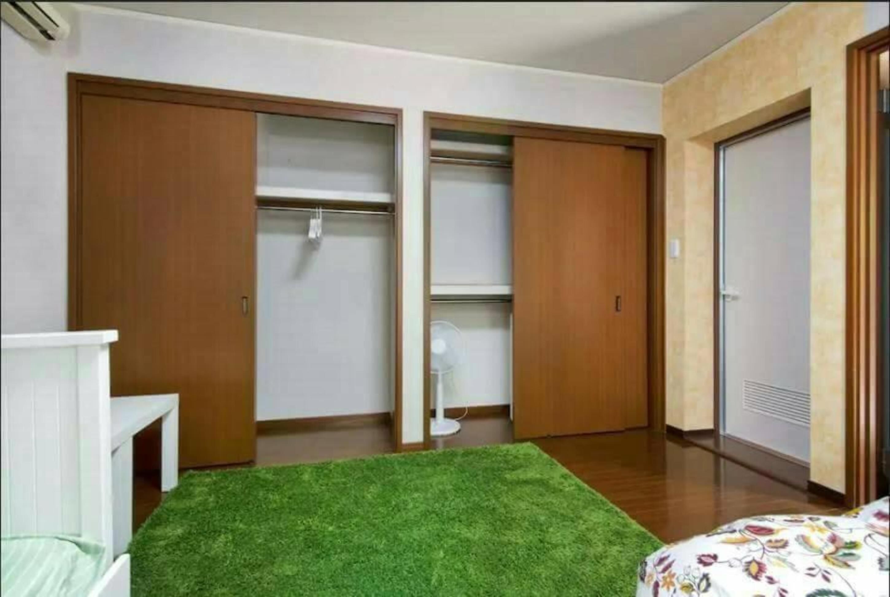A bedroom with a green artificial grass rug, wooden wardrobes, a white bedside table, and a bed with a floral bedspread.