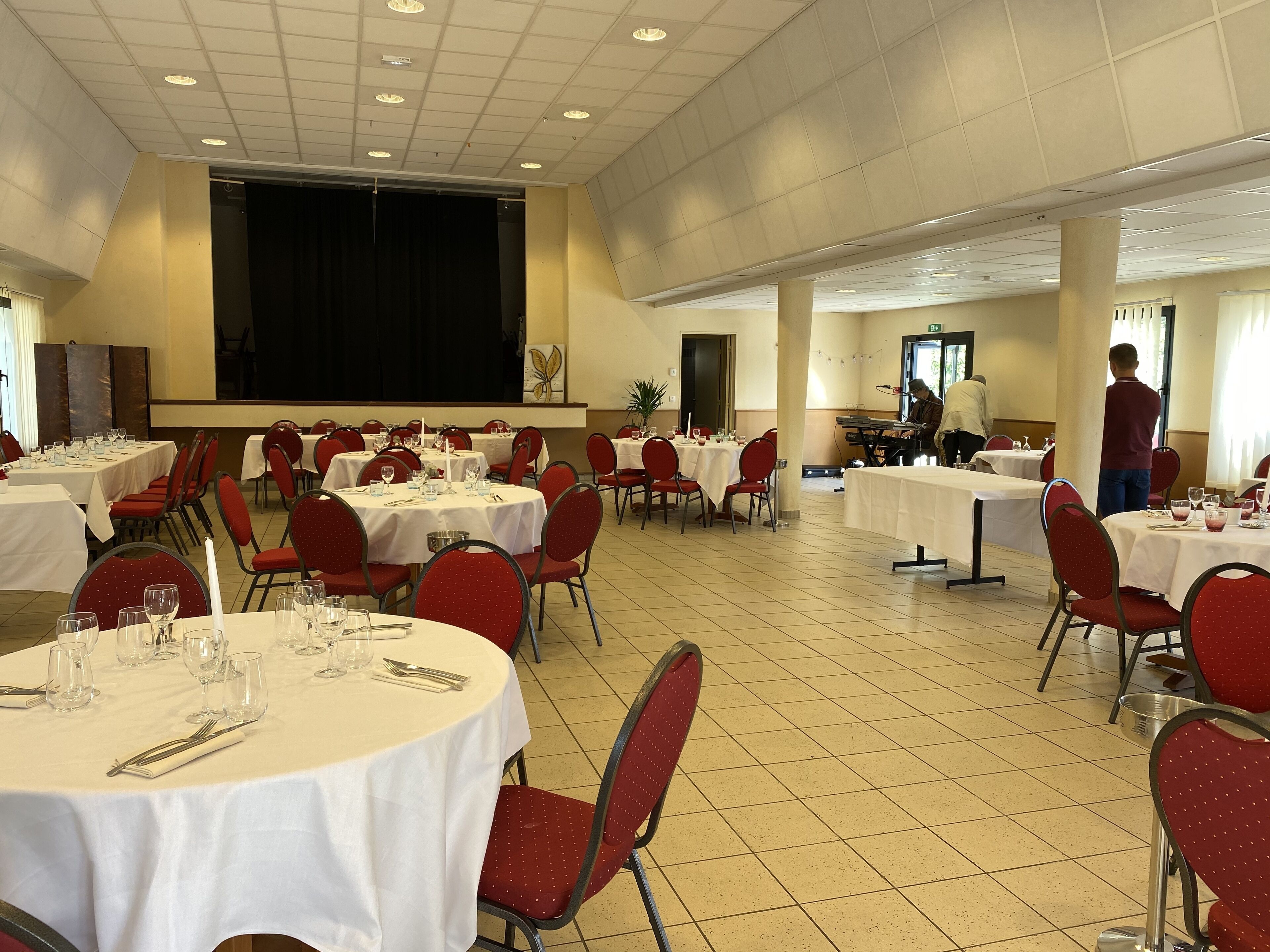 Een feestzaal met ronde tafels voor een diner, rode stoelen en een podium met een scherm.