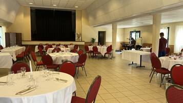 Een feestzaal met ronde tafels voor een diner, rode stoelen en een podium met een scherm.