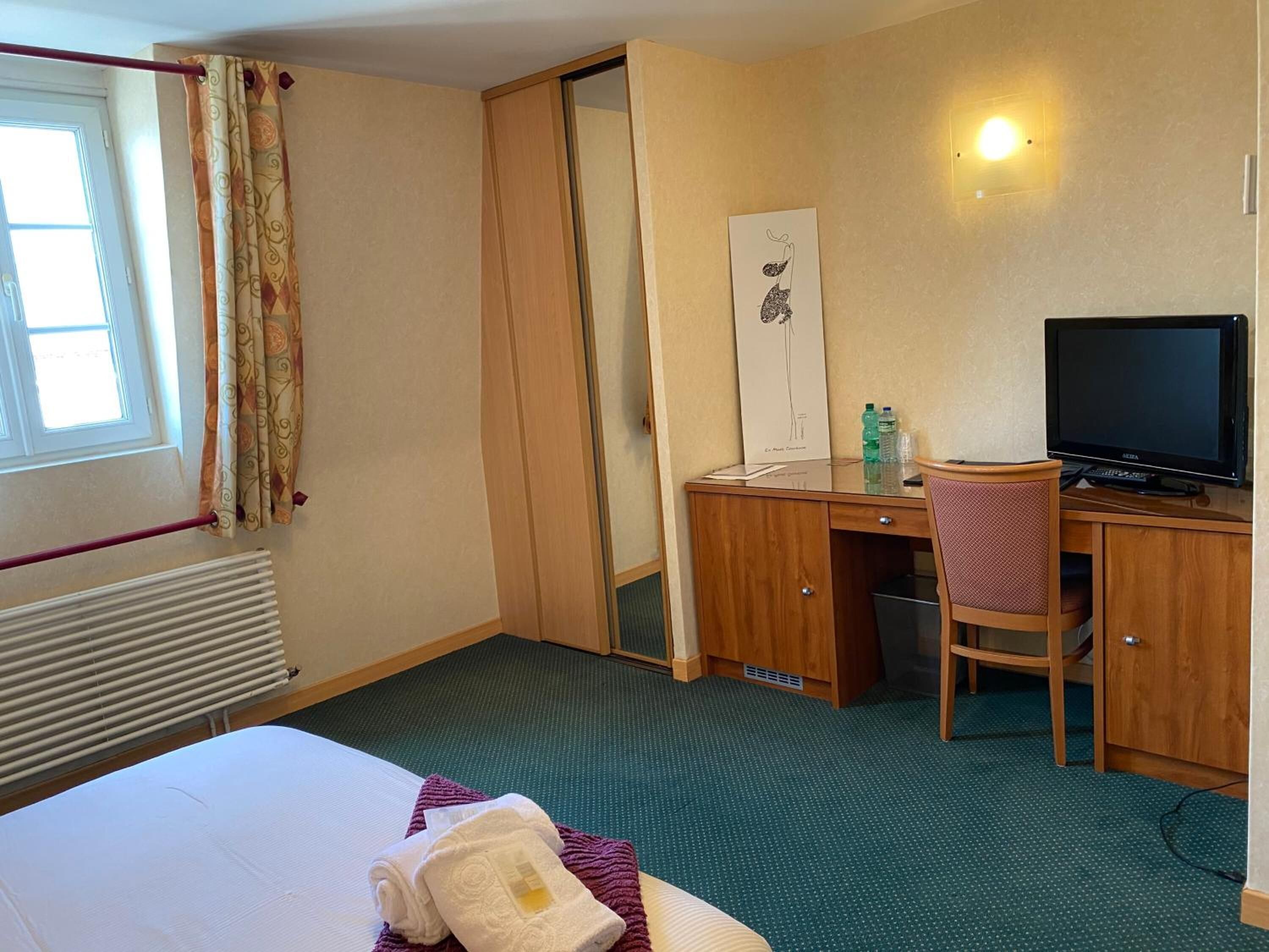 A hotel room with a bed, a desk with a computer and a chair, a television, and a window with curtains.