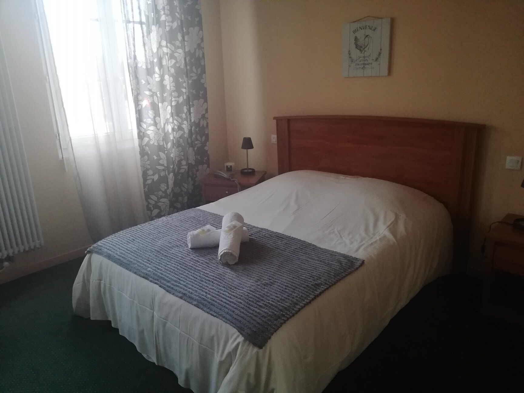 A hotel room with a bed, a nightstand, a lamp, a window with curtains, and a welcome sign on the wall.