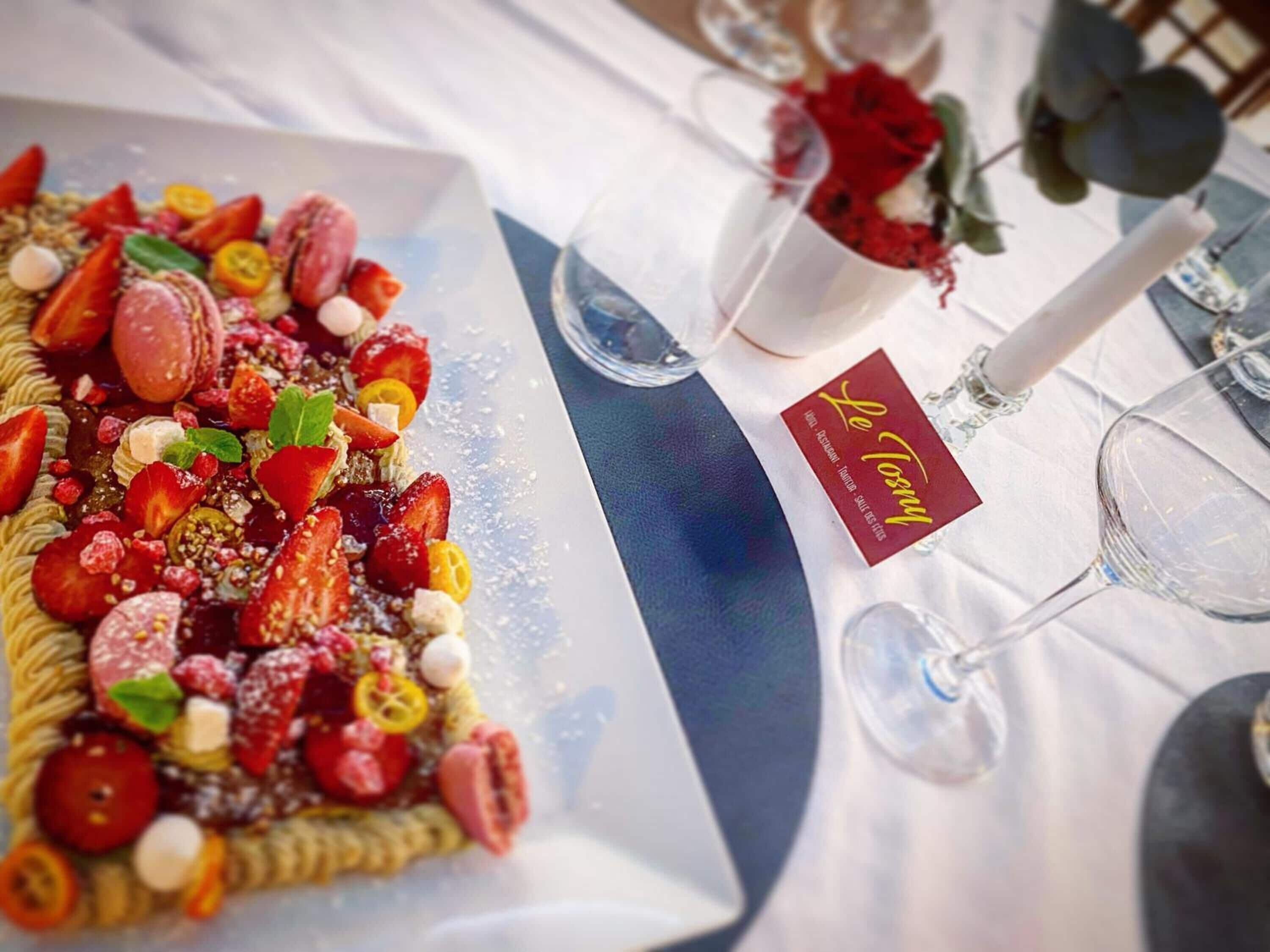 Un plato de postres con fresas, macarons y otras frutas, sobre una mesa con mantel blanco y una rosa en un florero.