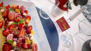 Un plato de postres con fresas, macarons y otras frutas, sobre una mesa con mantel blanco y una rosa en un florero.