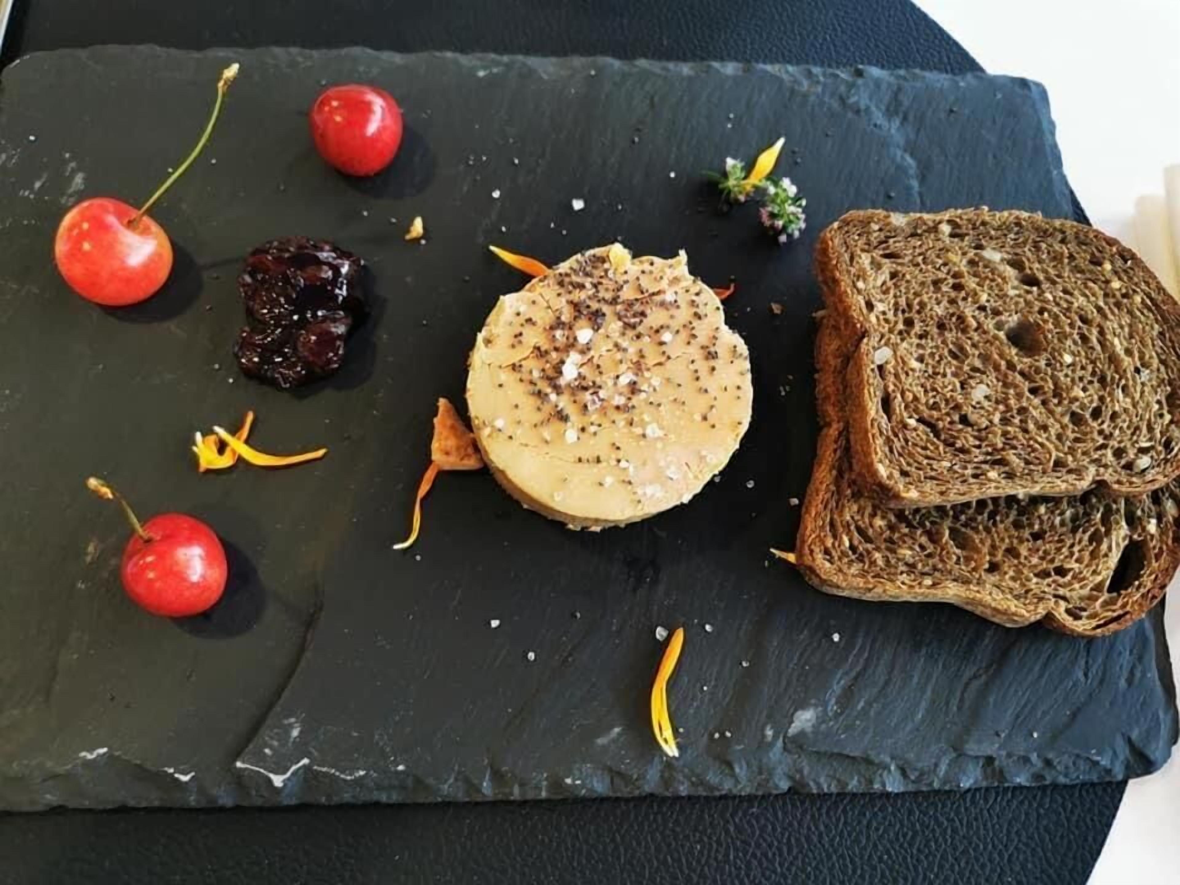 Een grijze serveerplank met een ronde kaas, gegarneerd met kruiden, kersen, gedroogd fruit en een plak donker brood.