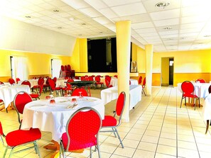 Un salón de banquetes con varias mesas redondas, sillas rojas y manteles blancos.