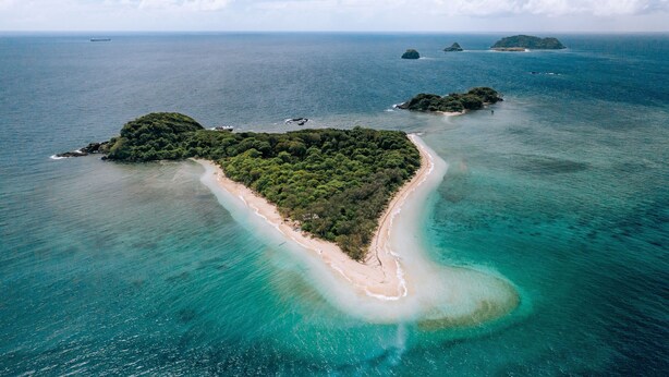 Luchtbeeld van een tropisch eiland met een smal strand, omgeven door turquoise water en weelderige begroeiing.