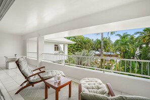 A balcony with a white railing, a cushioned chair, a wooden table, and a view of palm trees and a building. - Belle Escapes - Oceanview Suite Alamanda Beachfront Resort "88" (Palm Cove)