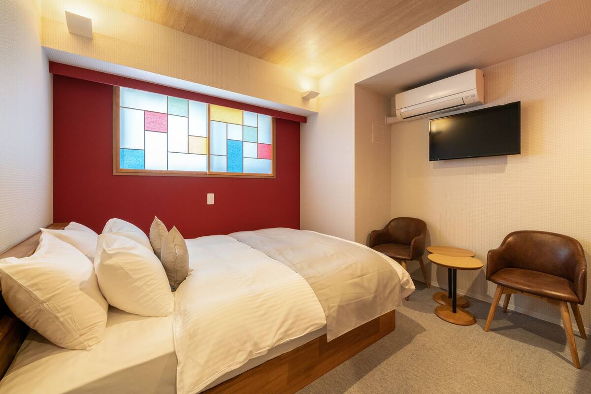 A hotel room with a bed, a small round table, two chairs, a TV, and a window with colorful panes.