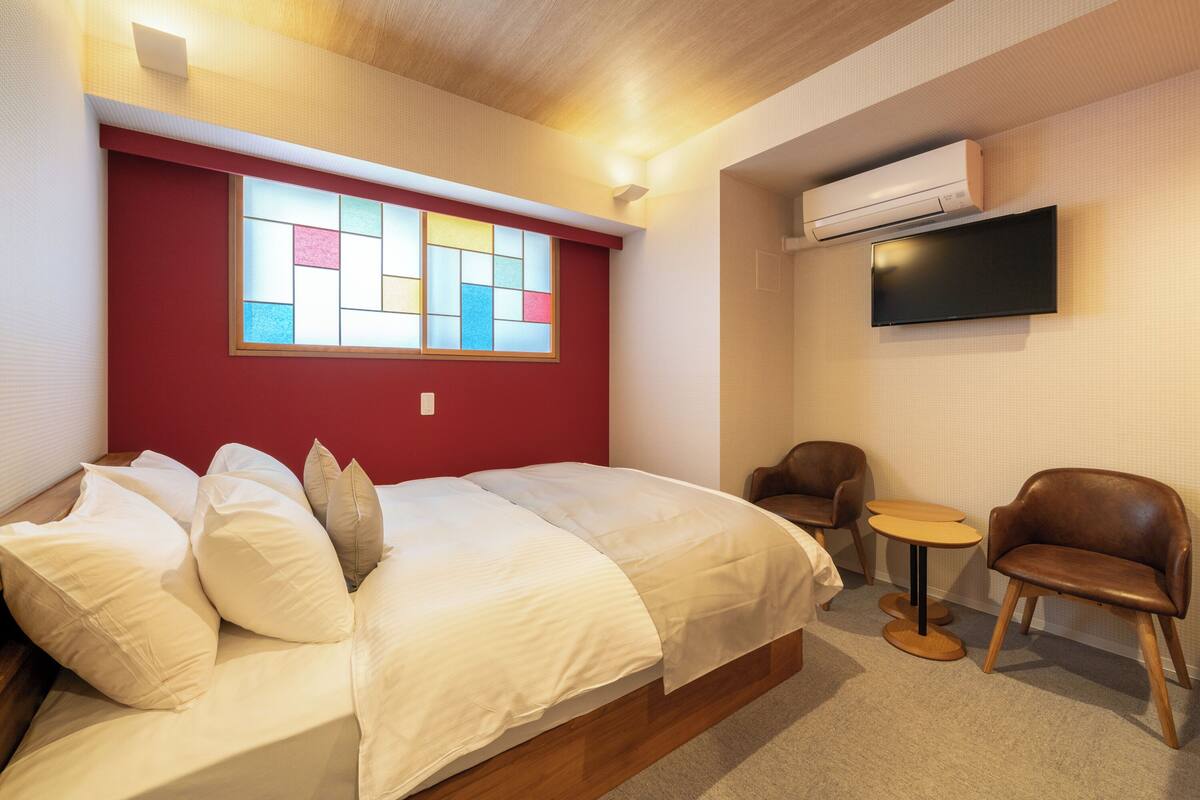A hotel room with a bed, a small round table, two chairs, a TV, and a window with colorful panes.