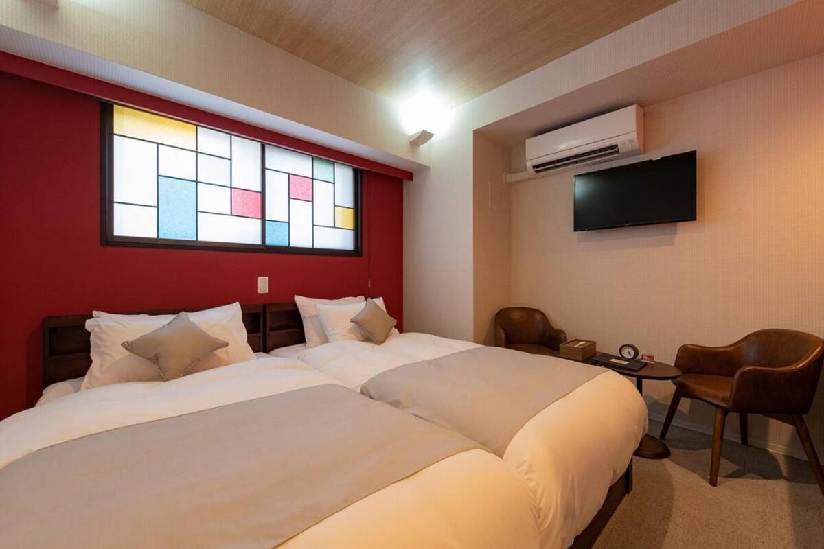 A hotel room with two beds, a TV, an air conditioner, a small table, and a chair.