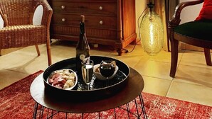 Ein gemütliches Wohnzimmer mit einer Holzkommode, Korbstühlen, einem Esstisch mit einer Platte voller Speisen, einer Flasche und einer Lampe.
