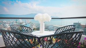 A balcony with a round table and chairs, a white vase with flowers, and a cityscape view. - Crystal Le Apartment Danang (Da Nang)