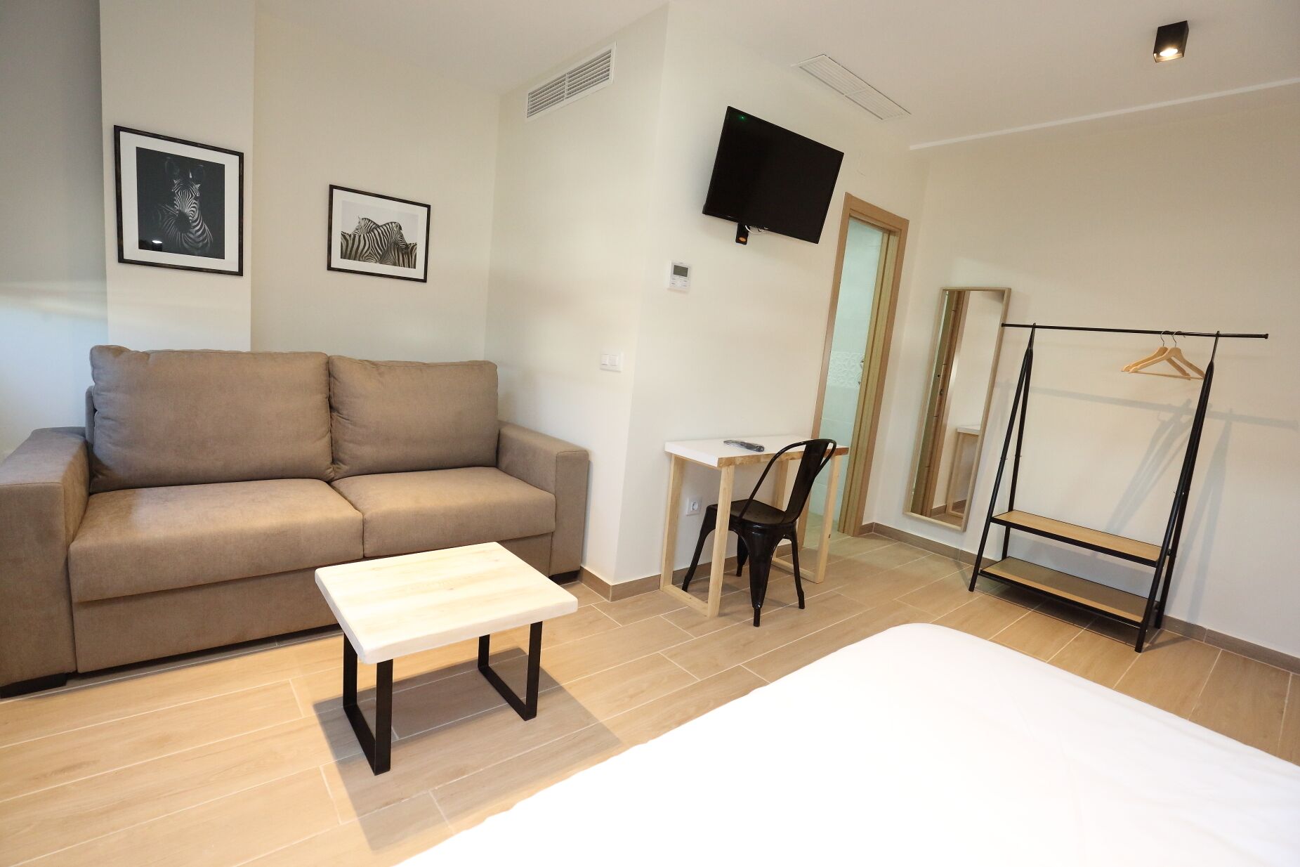 Ein modernes Zimmer mit einem beigefarbenen Sofa, einem kleinen weißen Tisch, einem schwarzen Sessel, einem an der Wand montierten Fernseher und einem Kleiderständer.