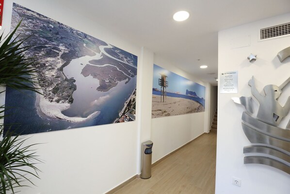 A hallway with a large mural of a coastal landscape, a reception desk, and a modern sculpture. - Hostal Palmones (Los Barrios)