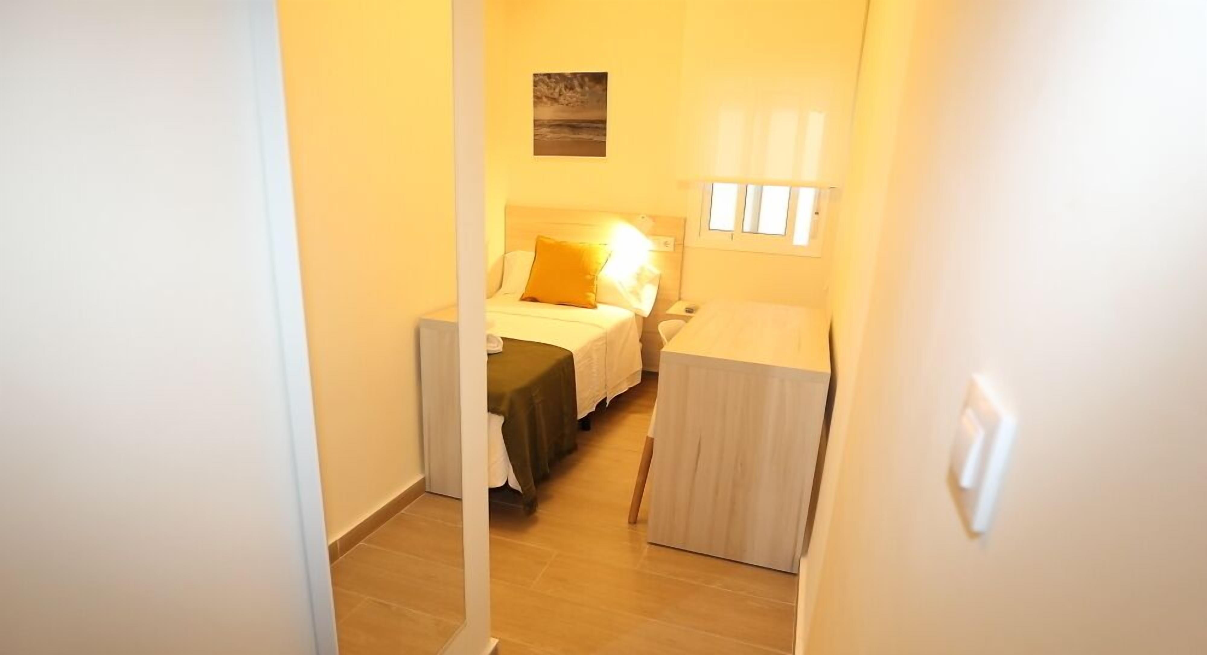 A narrow hallway leading to a bedroom with a bed, a bedside table, and a window.