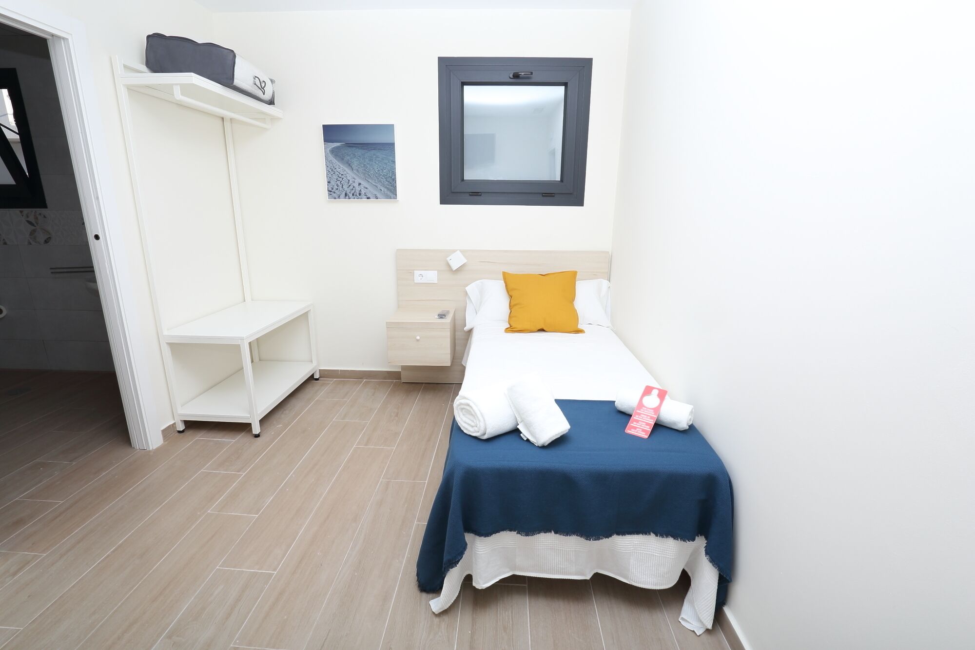 A neatly arranged bedroom with a bed, a bedside table, a nightstand, a shelf with a bag, and a picture on the wall.