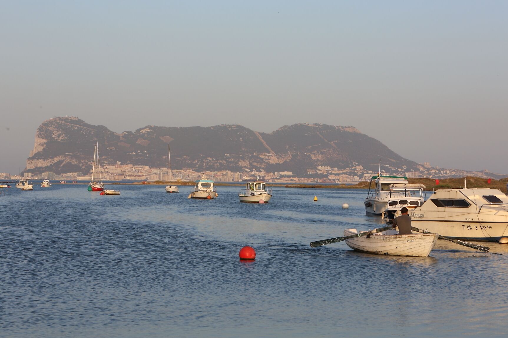 Un port de plaisance où sont amarrés plusieurs bateaux, une montagne imposante en arrière-plan et une eau calme.