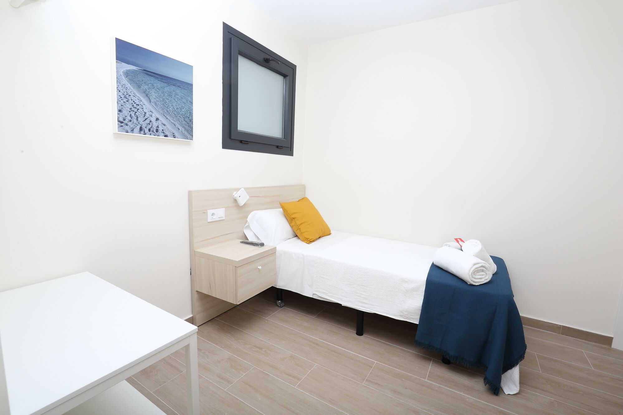 Une chambre avec un lit simple, une tête de lit en bois, une table de chevet, un bureau blanc et une couverture bleue sur le lit.