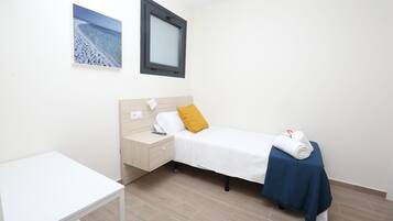 Une chambre avec un lit simple, une tête de lit en bois, une table de chevet, un bureau blanc et une couverture bleue sur le lit.