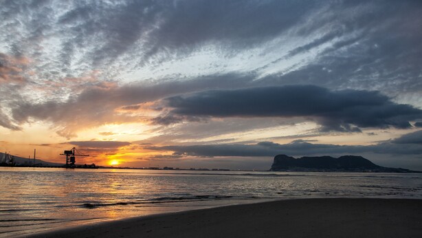 Un atardecer sobre una playa con estructuras industriales al fondo.