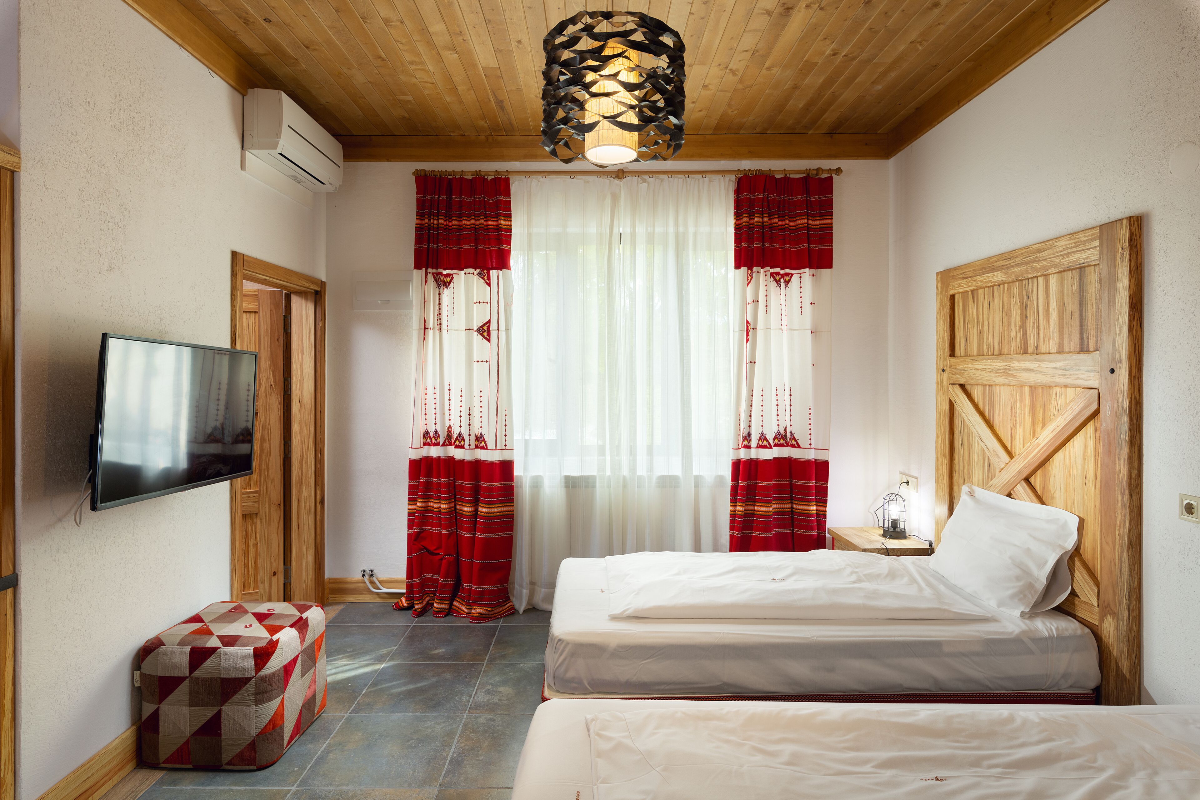 Un dormitorio con techo de madera, televisión de pantalla plana, dos camas, un banco y cortinas.