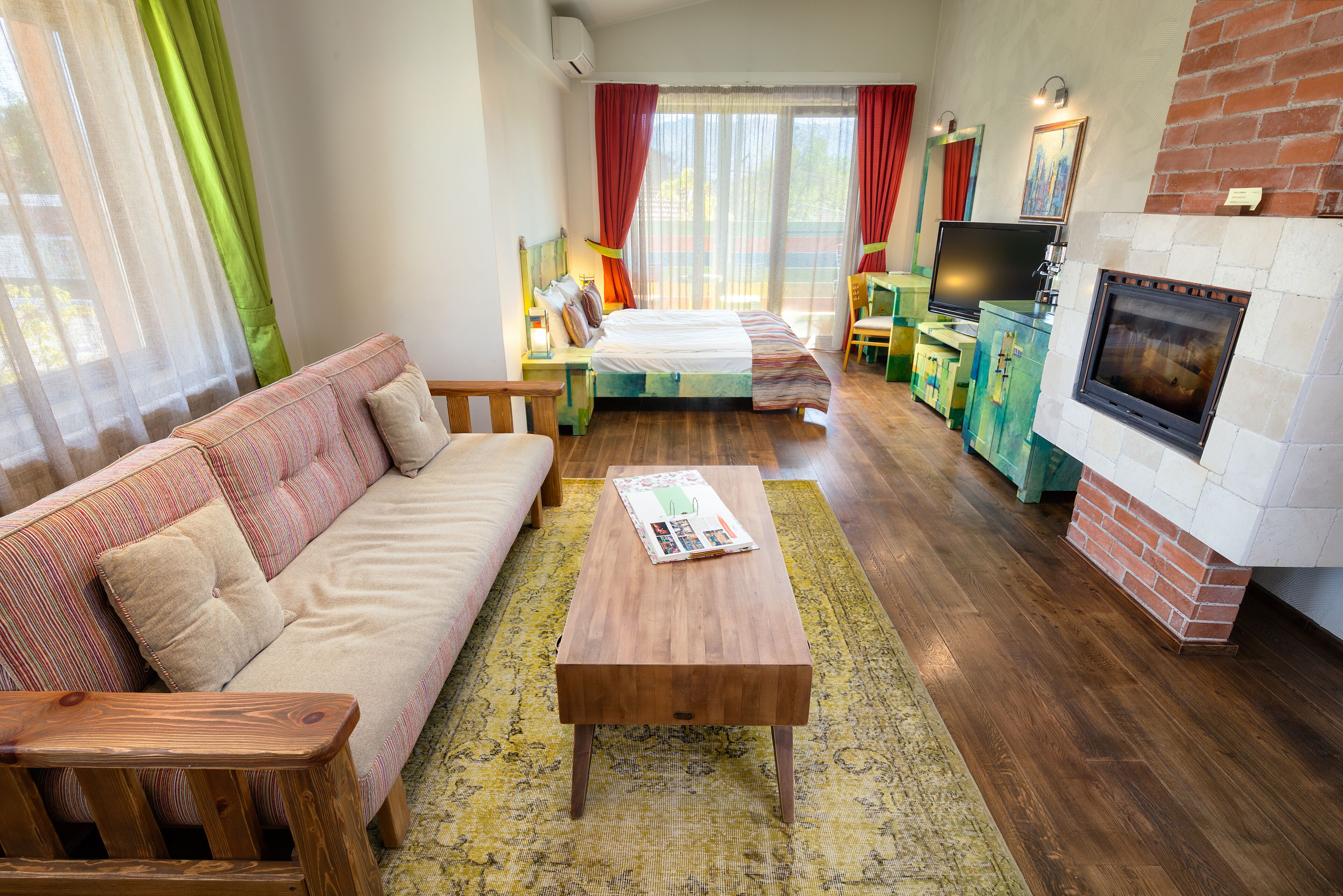 A hotel room with a sofa, a wooden coffee table, a bed, a TV, a fireplace, and a desk with a chair.