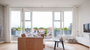 A spacious living room with a large window, a sofa, a small table, and a TV mounted on the wall. - Luxurious rooftop penthouse with rooftop terrace, a balcony, bath, duo shower (Groningen)