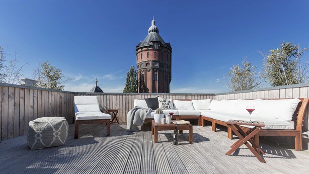 A rooftop terrace with a wooden deck, a white sofa set, and a view of a historic tower in the background. - Luxurious rooftop penthouse with rooftop terrace, a balcony, bath, duo shower (Groningen)