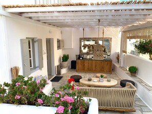 A sunlit outdoor seating area with a wooden table, wicker furniture, and potted plants. - Island Mykonos Suites & Studios (Mykonos)