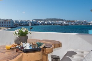 A wooden table with a potted plant, a croissant, sunglasses, a book, and a glass of orange juice on a terrace overlooking a coastal town with white buildings and blue waters. - Island Mykonos Suites & Studios (Mykonos)