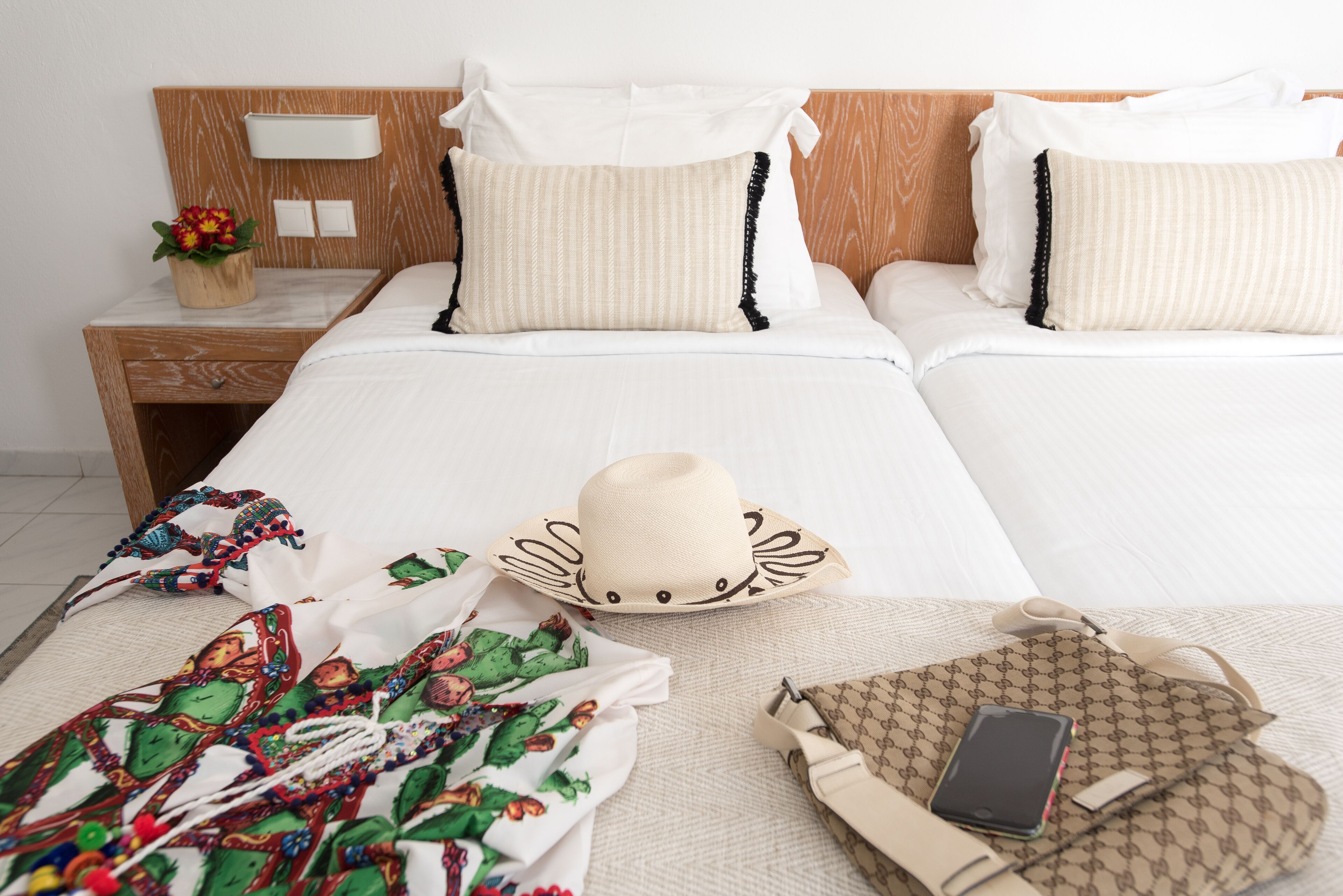 A hotel room with two beds, a nightstand with a lamp, a decorative plant, and a suitcase on the floor.